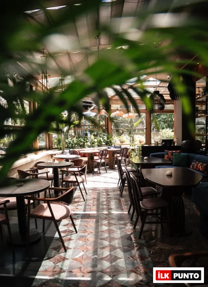 Doğal ışıkla aydınlanmış, bol bitki örtüsüyle çevrili şık bir kafe veya restoranın iç mekanı, desenli mermer zemini ve modern masa sandalye düzenlemeleriyle sıcak bir atmosfer sunarken, ön plandaki yemyeşil yapraklar derinlik ve tazelik katıyor.