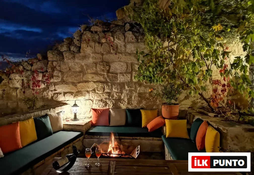 Kapadokya'da ziyaret edilebilecek en iyi mekanlardan 4 Oda Cave Hotel'in taş duvarlar arasına kurulmuş, ateş çukuru ve renkli minderlerle sıcak bir atmosfer sunan akşam ambiyansında, şarap kadehlerinin bulunduğu ahşap bir masa dikkat çekiyor.