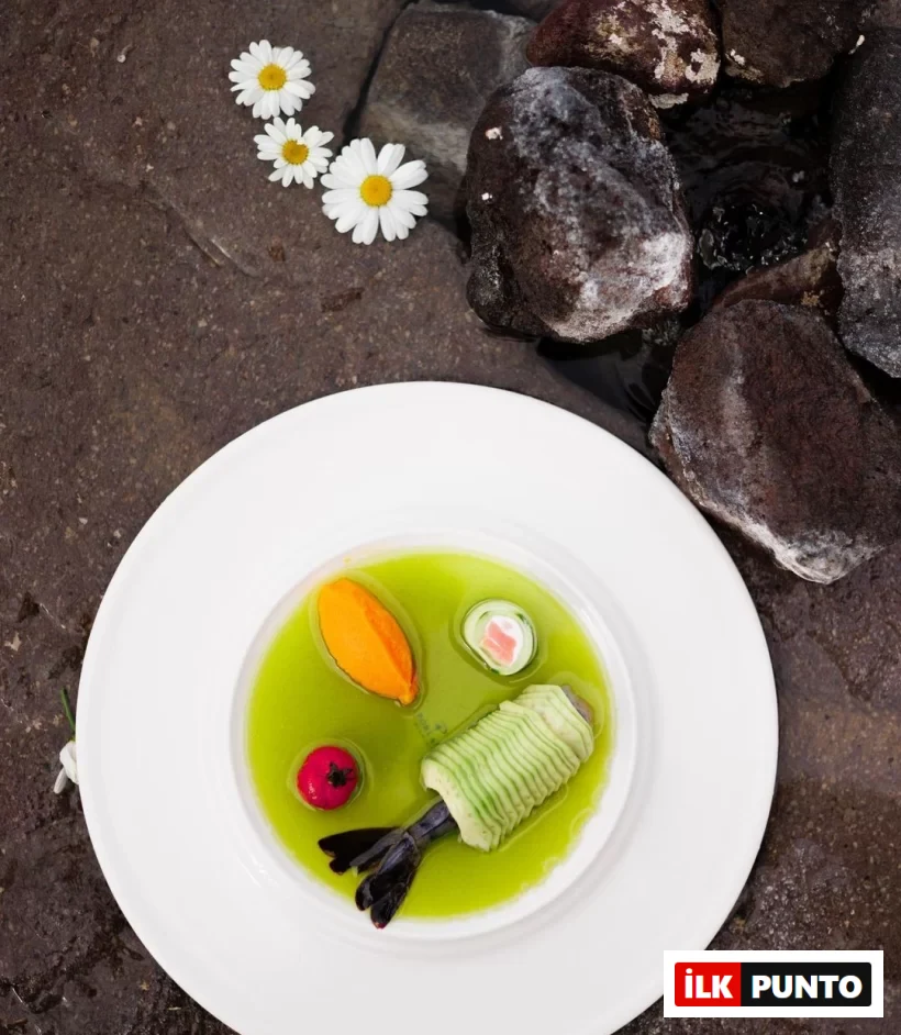 Kapadokya'da ziyaret edilebilecek gastronomik mekanların birinde servis edilen, açık yeşil bir sosun içinde özenle sunulan, ince dilimlenmiş salatalıkla sarılı büyük bir karidesin yanı sıra turuncu bir püre ve renkli garnitürlerle süslenmiş, arka planda ıslak koyu renkli taşlar ve papatyaların görüldüğü zarif bir tabak.