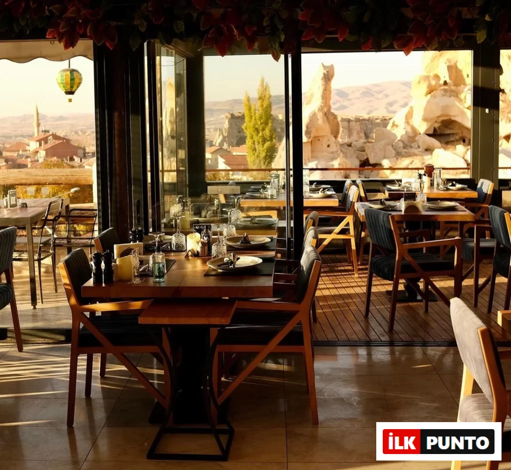 Kapadokya'da bulunan bu şık restoranın geniş pencerelerinden, masa düzenlemeleri gün batımının sıcak ışıklarıyla aydınlanırken, arka planda Çavuşin Kalesi'nin etkileyici manzarası, peribacaları ve uzakta yükselen renkli bir sıcak hava balonuyla birlikte unutulmaz bir akşam yemeği deneyimine davet eden atmosferi yansıtılıyor.