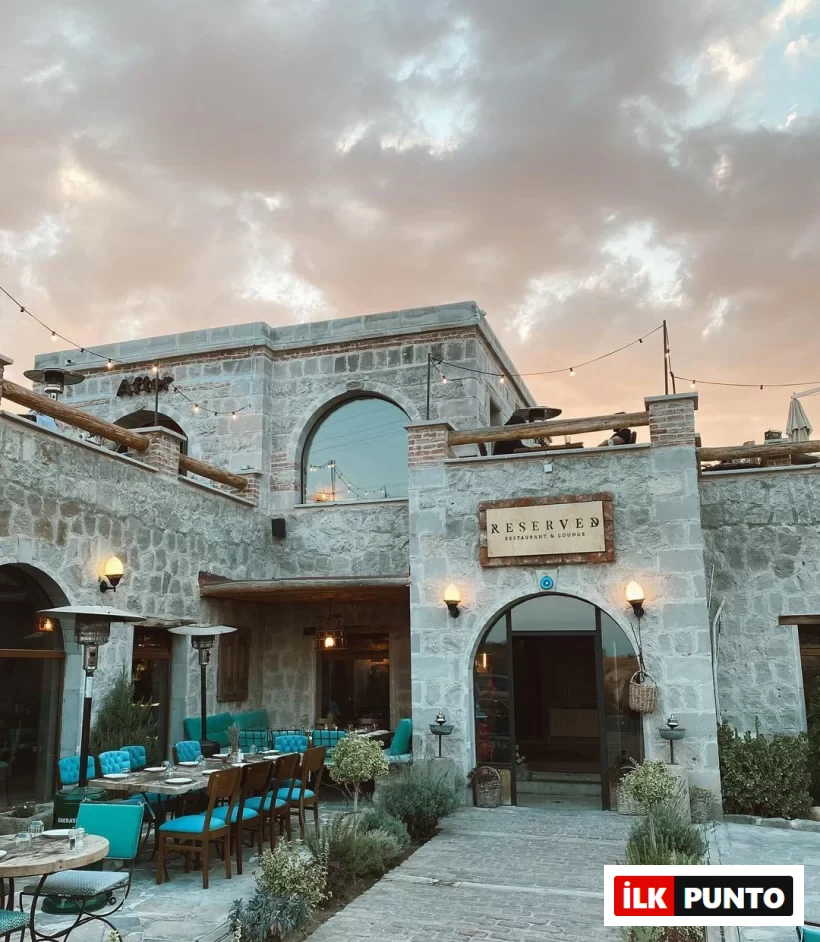 Reserved Restaurant & Lounge'ın Kapadokya'ya özgü taş mimarisiyle inşa edilmiş dış cephesi, gün batımının yumuşak ışıkları altında turkuaz sandalyeleri ve ahşap masalarıyla açık hava oturma alanını, asma ışıklar ve bitkilerle bezeli davetkar bir atmosferde sergiliyor.
