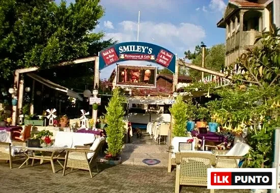 Smiley's Restaurant & Cafe'nin Arnavut kaldırımlı bir avluda yer alan, beyaz örtülü masalar ve hasır sandalyelerle dolu, yemyeşil ağaçlar ve sarı çiçekli bitkilerle çevrili, kısmen ahşap pergolalı açık hava yemek alanı gösterilmektedir; yukarıda ise balık şeklindeki tabelada restoranın balık, biftek ve daha fazlası sunduğu vurgulanmaktadır.