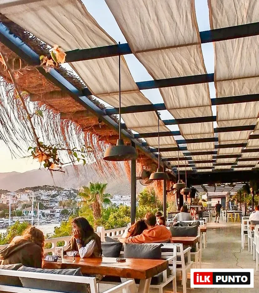 Kaş'ın büyüleyici manzarasına nazır bir açık hava restoranında, bej renkli kumaş tenteler ve sarkıt aydınlatmaların altında, ahşap masalarda oturan misafirler, gün batımının yumuşak ışıklarıyla aydınlanan denizi ve tepeleri izleyerek sakin bir akşamın keyfini çıkarıyor.