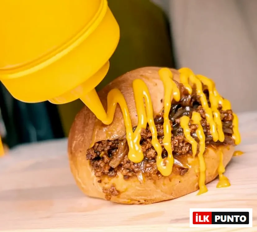 Açık renkli ahşap bir servis tahtası üzerinde duran, kıyma ve karamelize soğanlarla zengin bir şekilde doldurulmuş iştah açıcı bir hot dog sandviçin üzerine, sarı bir sos şişesinden akıcı bir şekilde parlak sarı bir sosun, muhtemelen hardal veya eritilmiş peynir sosunun, özenle döküldüğü yakın bir çekim.