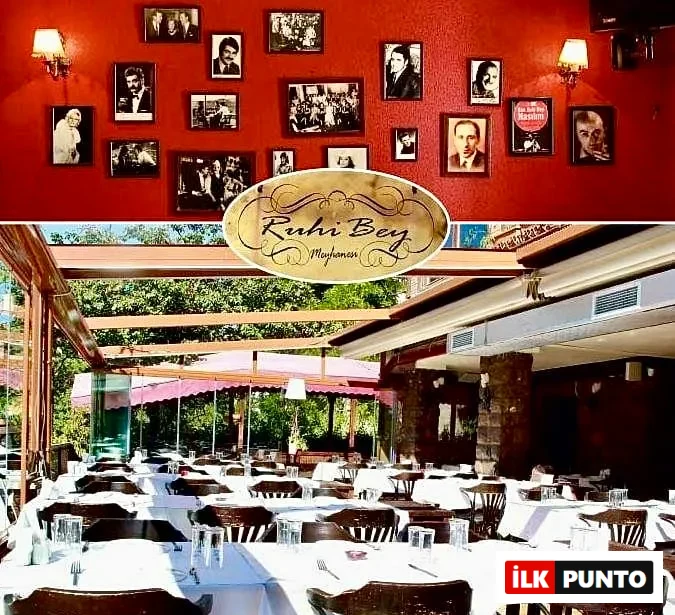 Ruhi Bey Meyhanesi'nin kırmızı duvarında asılı nostaljik siyah beyaz portre ve toplu an fotoğrafları ile beyaz örtülü masaların ve ahşap sandalyelerin dizili olduğu ferah dış mekan yemek alanı birlikte görülüyor.