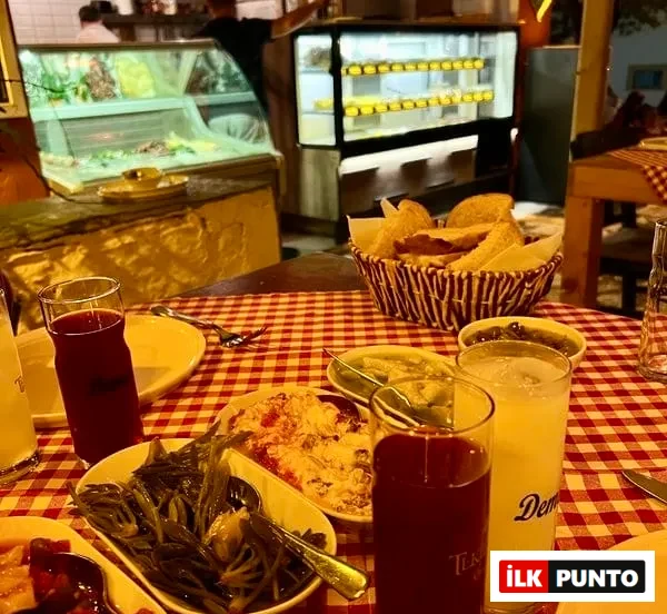 Kırmızı beyaz kareli bir masa örtüsü üzerinde, Demeti markalı buzlu rakı ve kırmızı bir içecek bardağının yanı sıra, deniz börülcesi, yoğurtlu ve domatesli mezelerin bulunduğu tabaklar ile bir ekmek sepeti yer alırken, arka planda bir restorana ait vitrinler ve hafifçe flu insanlar görülen, tipik bir Türk meyhanesi sofrası yansıması.