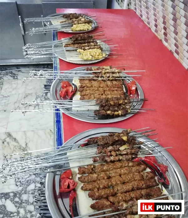 Kırmızı bir masa üzerinde yan yana dizilmiş metal tepsilerde, lavaş ekmeği üzerinde sunulan bolca Adana kebap, kuşbaşı et ve tavuk şiş kebap çeşitleri ile ızgara domates ve biberler, servise hazır bir şekilde bekliyor.