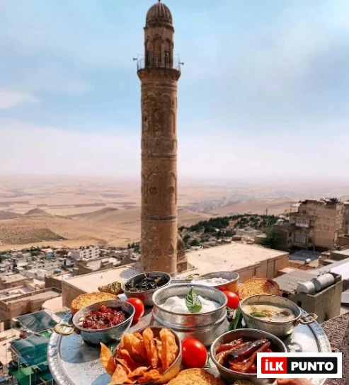 Gümüş bir tepside sunulan zeytin, yoğurtlu soslar, közlenmiş biber ve kızarmış patates gibi lezzetli atıştırmalıkların ön planda olduğu, arka planda ise tarihi Mardin'in eşsiz taş mimarisi, yüksek minaresi ve Mezopotamya Ovası'nın nefes kesen genel görünümüyle seyirlik bir manzara.