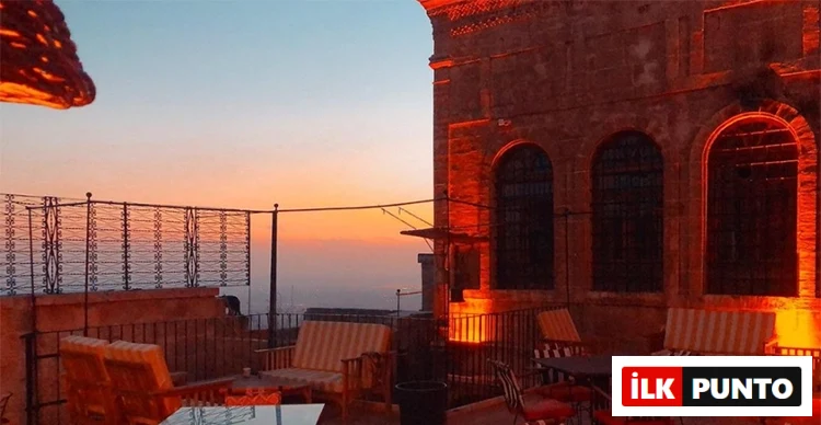 Gökyüzünün turuncu ve mavi tonlarına büründüğü gün batımı manzarasıyla, sıcak portakal rengi ışıklarla aydınlatılmış, kemerli pencerelere sahip tarihi bir taş binanın yanı başında yer alan şık bir terasta, rahat koltuk takımları uzaklara nazır keyifli bir dinlenme alanı sunuyor.