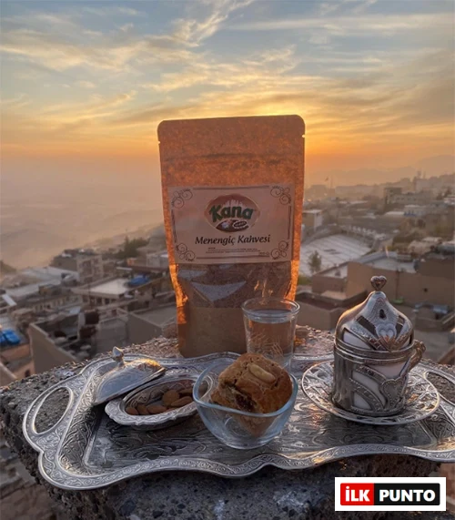 Güneş batımının altın renkleriyle parlayan, Mardin'in eşsiz tarihi şehir manzarasına hakim bir taş zeminde yer alan, üzerinde Kana Menengiç Kahvesi paketi, yanında bademli kurabiye, bir bardak su ve zarif işlemeli gümüş bir kahve fincanının bulunduğu geleneksel bir tepsi, huzurlu bir anı yansıtıyor.
