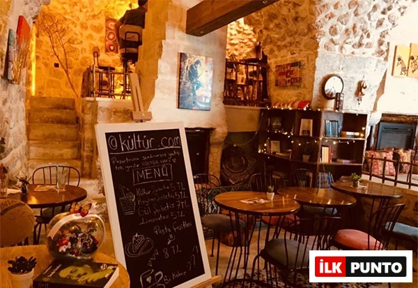 Taş duvarlara sahip, loş ışıklı, samimi bir kültür kafe iç mekanı; ön planda bir menü tahtası, arka planda yuvarlak masalar, sandalyeler, raflarda ve masalarda kitaplar ile duvarlarda sanat eserlerinin bulunduğu, huzurlu bir ortam sunuyor.