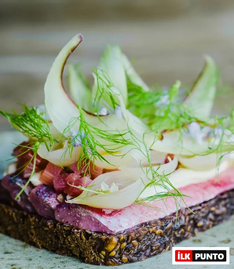 Selma restoranından enfes bir örnek olan bu smørrebrød, koyu renkli, tanecikli çavdar ekmeği üzerinde pembe tonlarda marine edilmiş ringa balığı dilimleri, kremsi beyaz sos, zarifçe kıvrılmış rezene dilimleri ve bolca taze dereotuyla süslenmiş, küçük mor çiçeklerle detaylandırılmış eşsiz bir Kopenhag lezzetini gözler önüne seriyor.