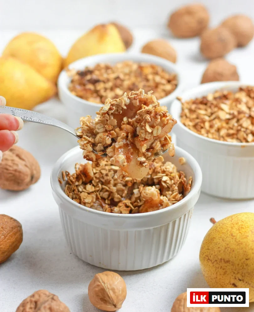 Bir kaşık dolusu yulaflı armut crumble, beyaz bir fırın kabından kaldırılırken yakın çekimde görülüyor; arka planda bulanık duran diğer crumble kapları ve etrafa dağılmış taze armutlar ile cevizler, bu lezzetli tatlının doğal ve besleyici içeriğini vurguluyor.