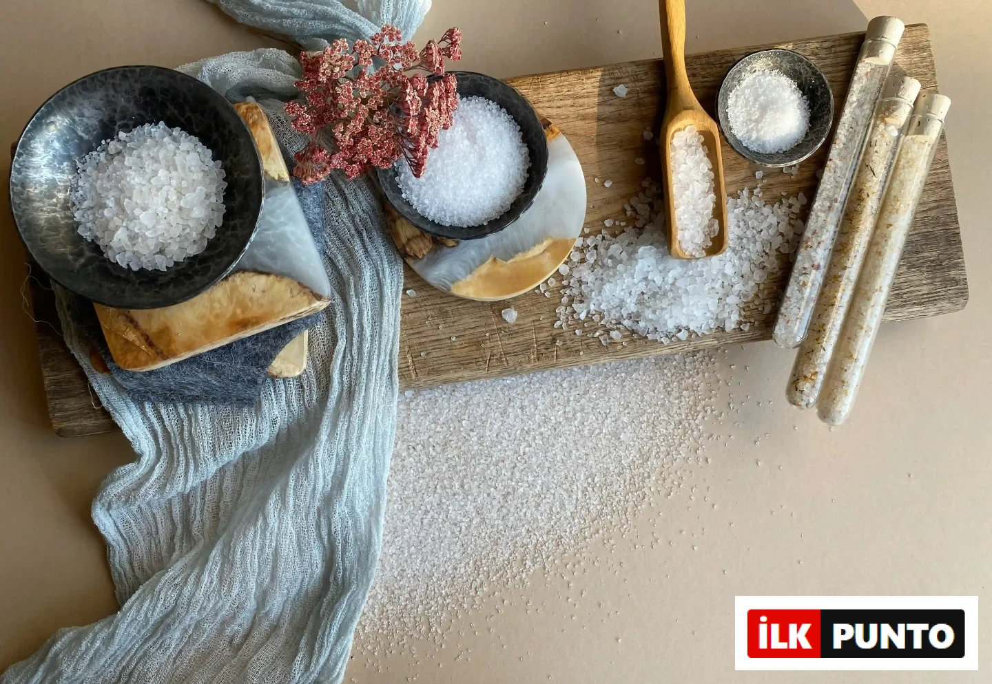 Ahşap bir kesme tahtası üzerinde, iki koyu renk kasede iri taneli, küçük bir gümüş kapta ve ahşap kaşıkta farklı tuz çeşitleri ile mantar tıpalı üç cam tüpteki aromalı tuzlar, açık mavi dokulu kumaş ve bej zemine dağılmış tuz kristalleriyle birlikte estetik bir düzenleme oluşturuyor.