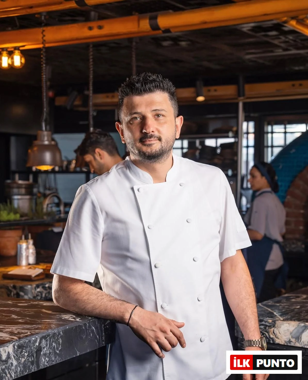 Şef Mevlüt Özkaya, beyaz aşçı ceketini giymiş, kalabalık bir restoran mutfağında koyu renkli mermer bir tezgaha yaslanarak kameraya doğrudan bakıyor; arkasında çalışanlar ve endüstriyel aydınlatmalar modern ve hareketli bir ortamı vurguluyor.