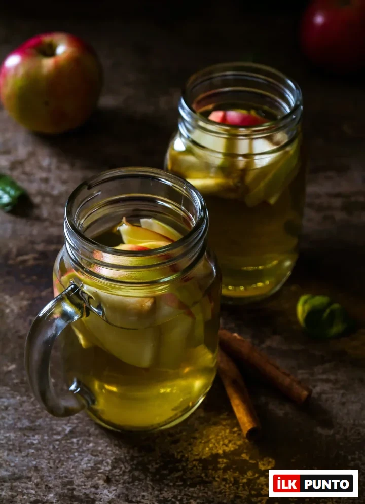Karanlık ahşap bir yüzey üzerinde, taze elma dilimleri ve iki tarçın çubuğuyla zenginleştirilmiş, sağlıklı bir elma sirkesi veya ferahlatıcı elma infüzyonu içeren iki cam kavanoz, sıcak bir atmosfer sunuyor.