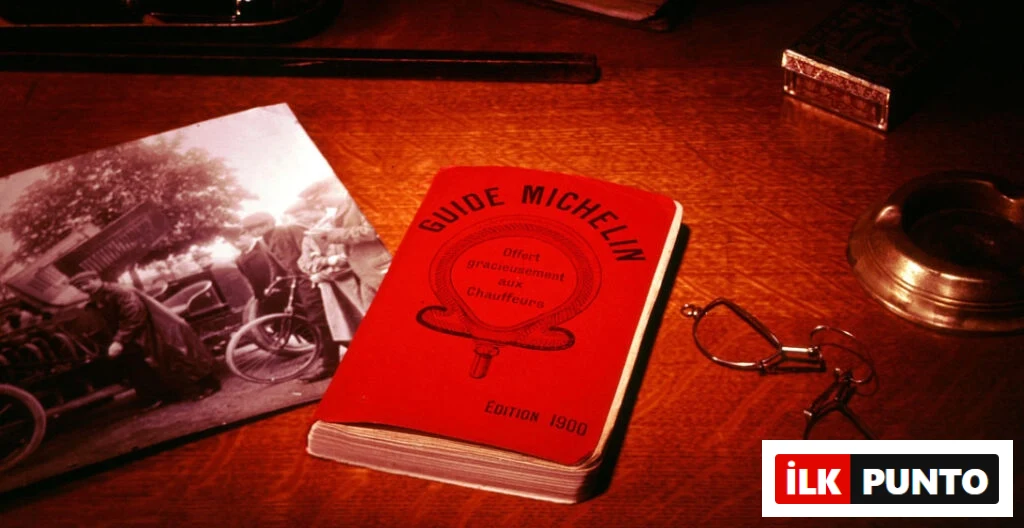 1900 yılına ait ilk Michelin Rehberi’nin kırmızı kapaklı baskısı, eski bir masa üzerinde gözlük ve siyah beyaz bir fotoğrafla birlikte sergileniyor.