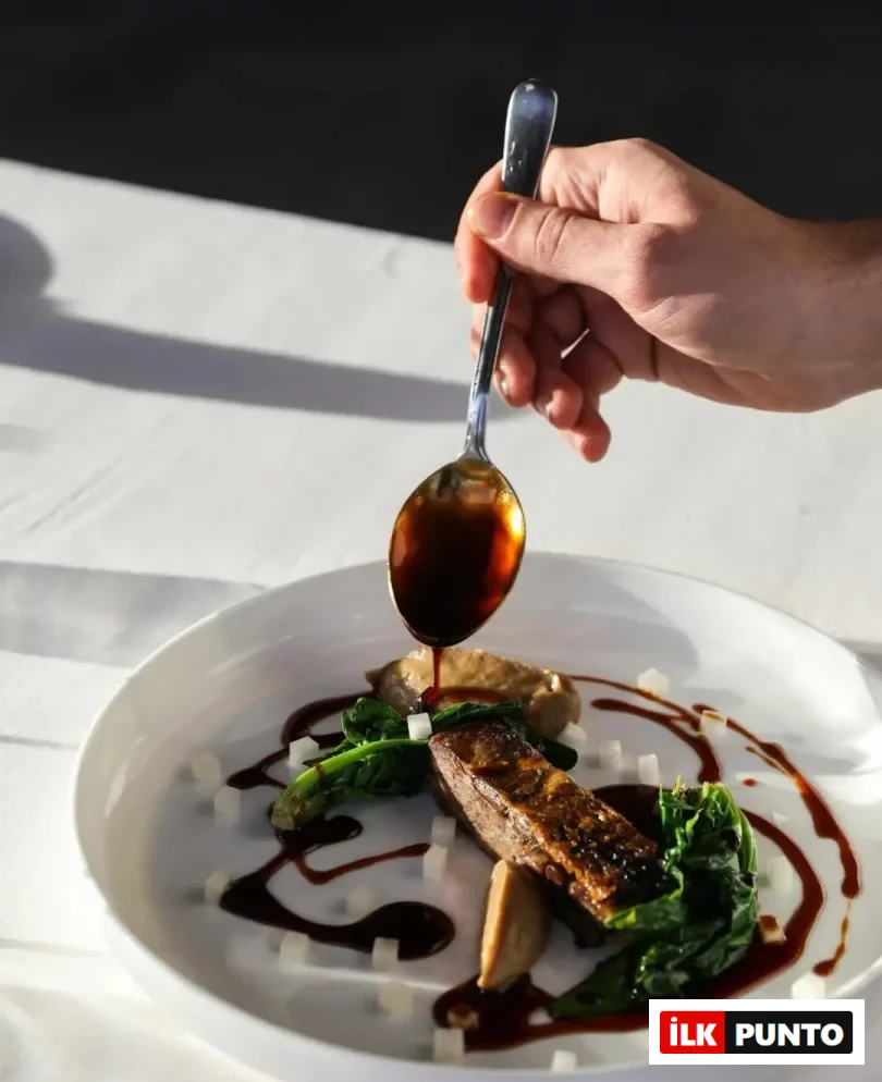 Muğla'nın Michelin Yıldızlı restoranlarından Maçakızı'nın zarif mutfak anlayışını yansıtan bu karede, bir elin kaşıkla koyu renkli sosu dikkatlice döktüğü, mühürlenmiş et, taze yeşillikler ve modern garnitürlerle sanatsal bir şekilde sunulmuş ana yemek tabağı görülüyor.
