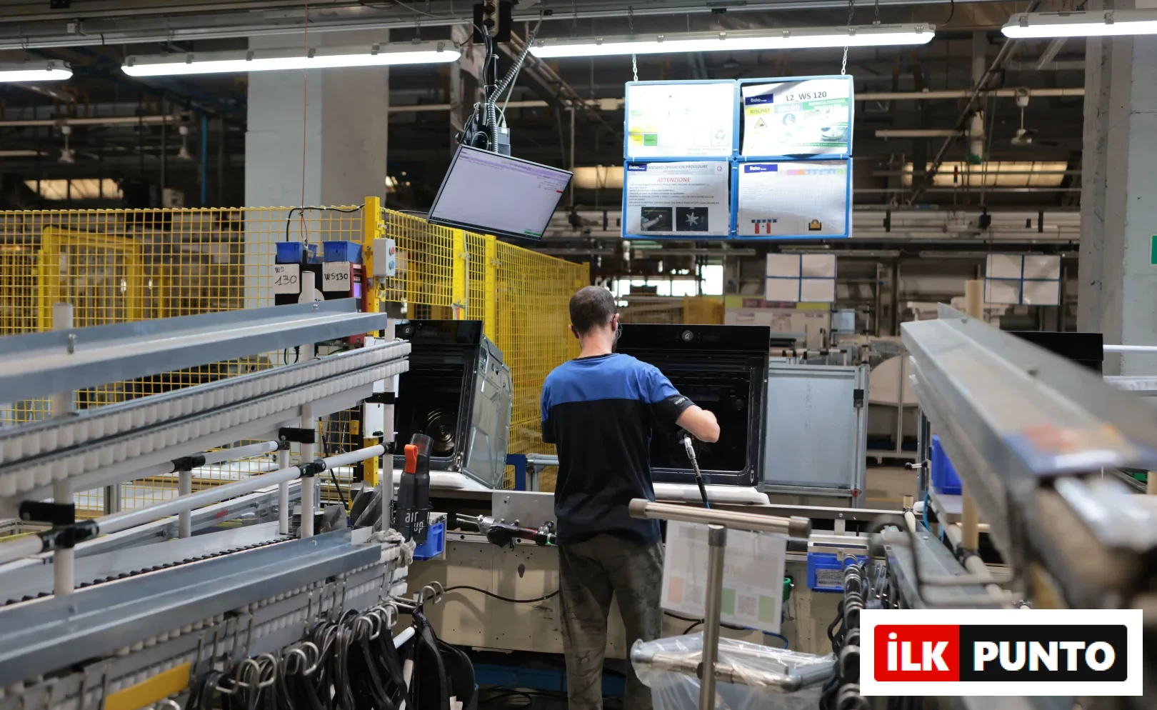 Beko Europe Cassinetta fabrikasında, montaj hattında bir fırının iç aksamlarını özenle birleştiren mavi-siyah tişörtlü bir çalışan, ankastre teknolojilerindeki üretim ayak izini gösteriyor; etrafında konveyör bantlar ve dijital ekranlarda enerji sınıfı bilgileri görülüyor.