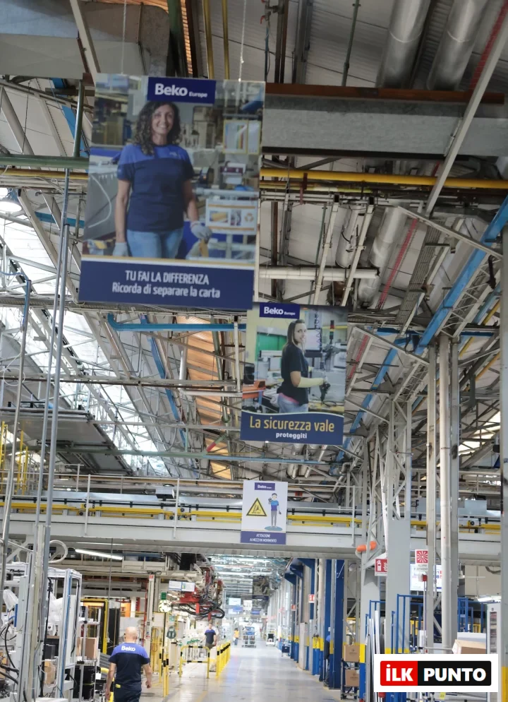 Beko Europe markalı iş kıyafetleri giyen çalışanların yürüdüğü, modern makineler ve İtalyanca güvenlik ile sürdürülebilirlik mesajları içeren afişlerle dolu geniş bir fabrika üretim hattı, Arçelik'in Avrupa'daki üretim ayak izi ve küresel rekabetteki stratejik gücünü temsil eden Cassinetta İşletmeleri'nden bir kesit sunuyor.