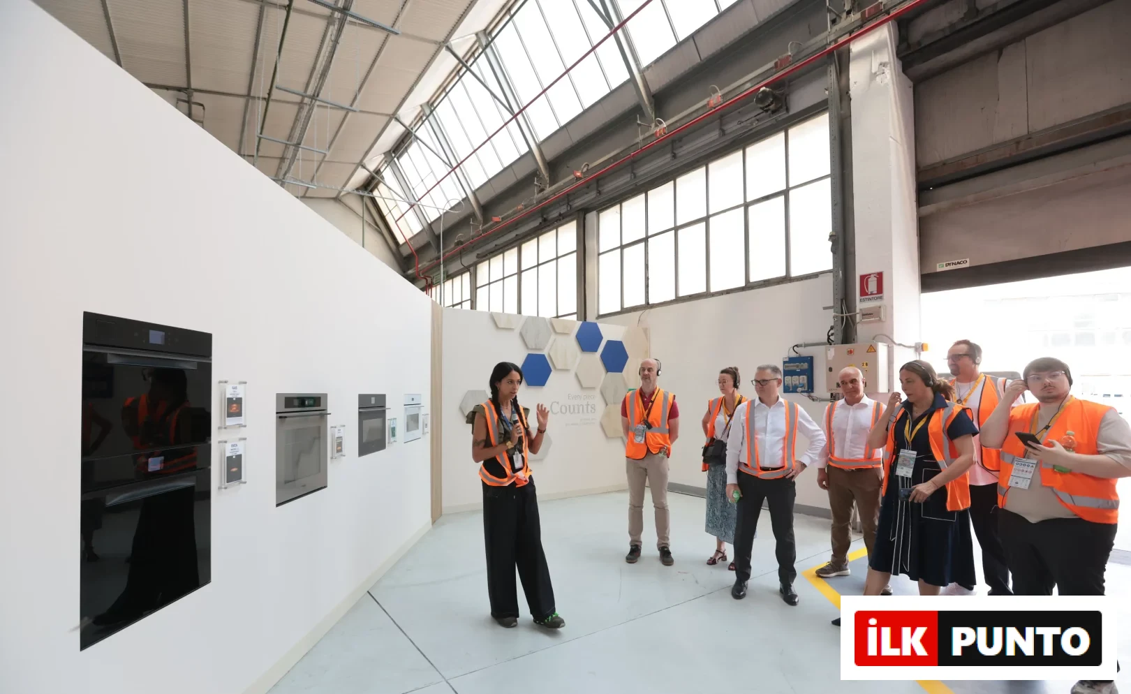 Kuzey İtalya’daki Beko Europe’un devasa Cassinetta İşletmeleri’nde düzenlenen basın gezisinde, turuncu yelekli bir grup, beyaz bir panelde sergilenen çeşitli ankastre fırın ve mutfak cihazlarını dinleyen turuncu yelekli bir konuşmacıyı takip ediyor, bu da şirketin marka stratejisi ve üretim ayak izini vurguluyor.