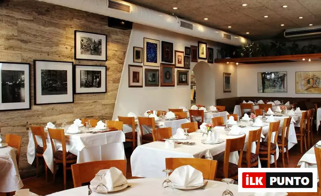 İlk Punto - Salvatore Cannetiello – Şeflerin Lezzet Rotaları: Şefin balık ve meze favorisi Tarabya'daki Kıyı Balık restoranının iç mekanı. Beyaz masa örtüleri, ahşap sandalyeler, özenle katlanmış peçetelerle kurulan masalar ve taş duvarları süsleyen çerçeveli fotoğraflar sıcak bir ortam sunuyor.