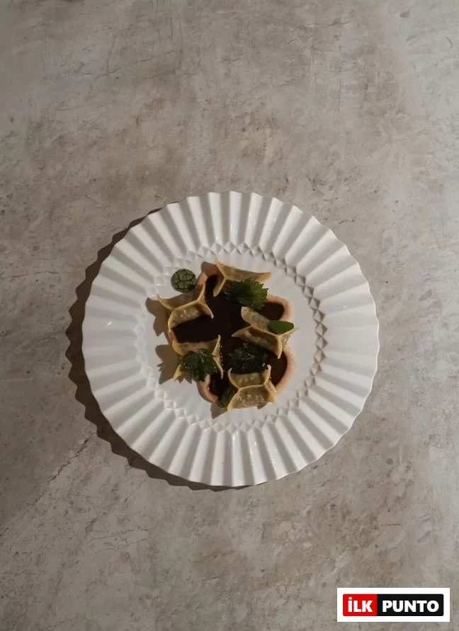 Beyaz, dalgalı kenarlı zarif bir tabakta sunulan, koyu soslu ve taze yeşilliklerle süslenmiş modern mantı benzeri yemek, Urla'nın yenilikçi gastronomi anlayışını ve özgün lezzetlerini vurguluyor.