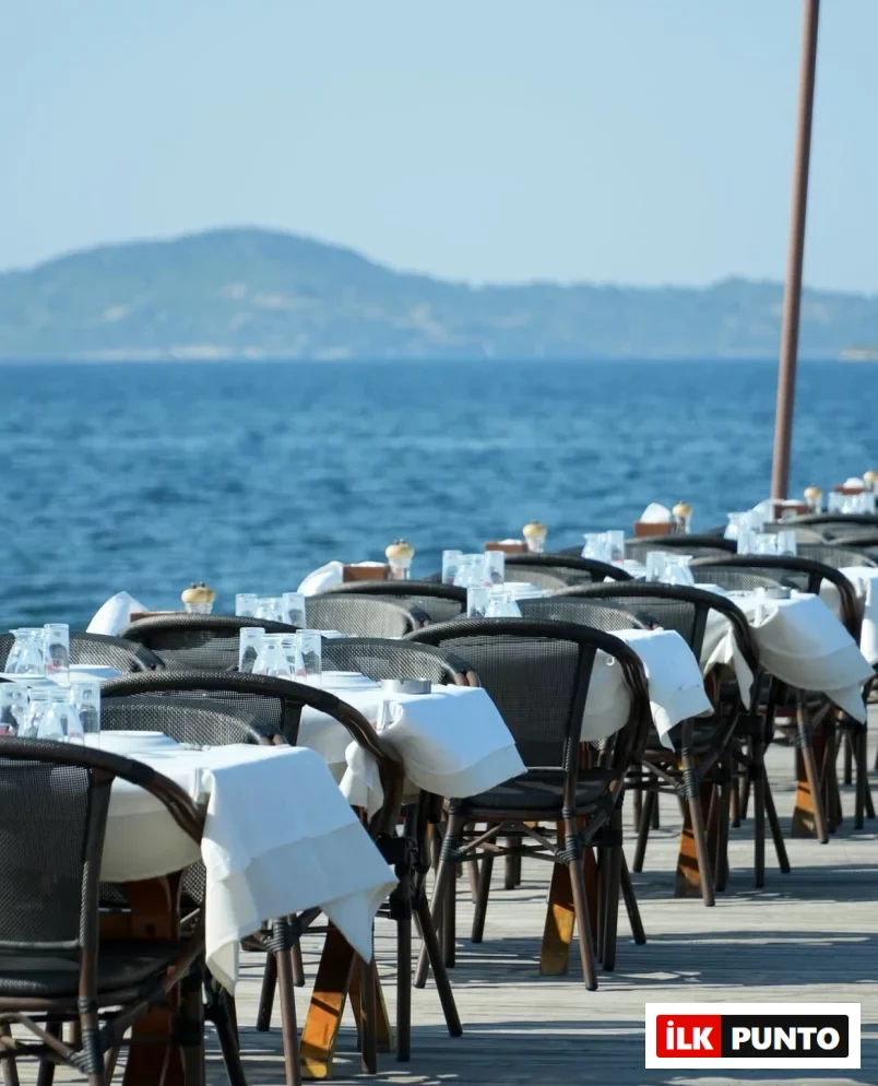 Urla sahilindeki bir işletmede, masmavi denize sıfır ahşap bir iskele üzerinde güneşin altında sıra sıra dizilmiş, beyaz örtülü, boş masa ve hasır sandalyeler ile uzakta belli belirsiz dağ silüetleri yer alıyor.
