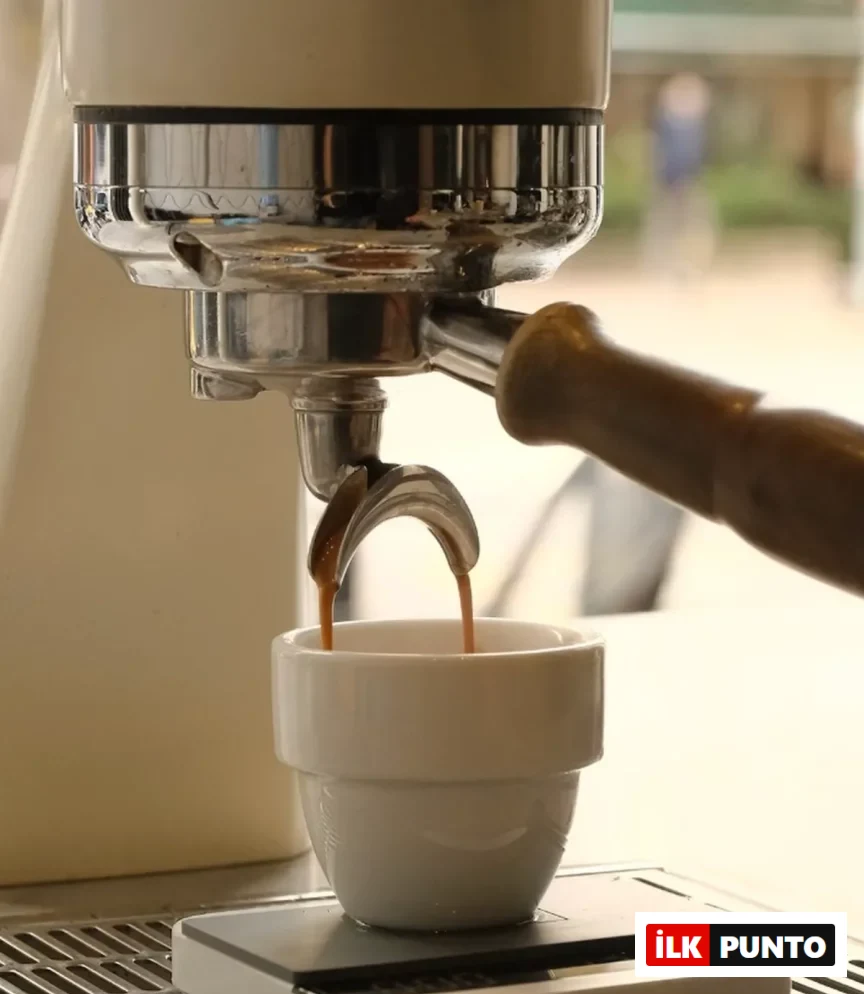 Bir espresso makinesinden portafiltrenin çift oluklu çıkışından küçük, şık beyaz bir fincana yavaşça akan koyu kahve, en iyi kahvecilerde deneyimleyebileceğiniz kaliteli kahve hazırlama sürecini canlı bir şekilde gösteriyor.