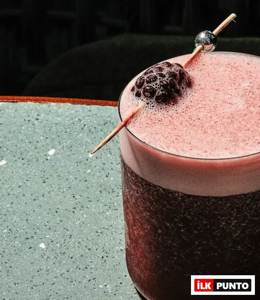 İstanbul'daki Manze restoranında sunulan, koyu kırmızı renkte, üzerinde pembe köpük tabakası bulunan ve ahşap çubuğa takılı parlak bir böğürtlen ile gümüş rengi bir boncukla zarifçe süslenmiş serinletici bir kokteylin yakından çekilmiş görüntüsü, yeşil benekli bir masanın üzerinde duruyor.