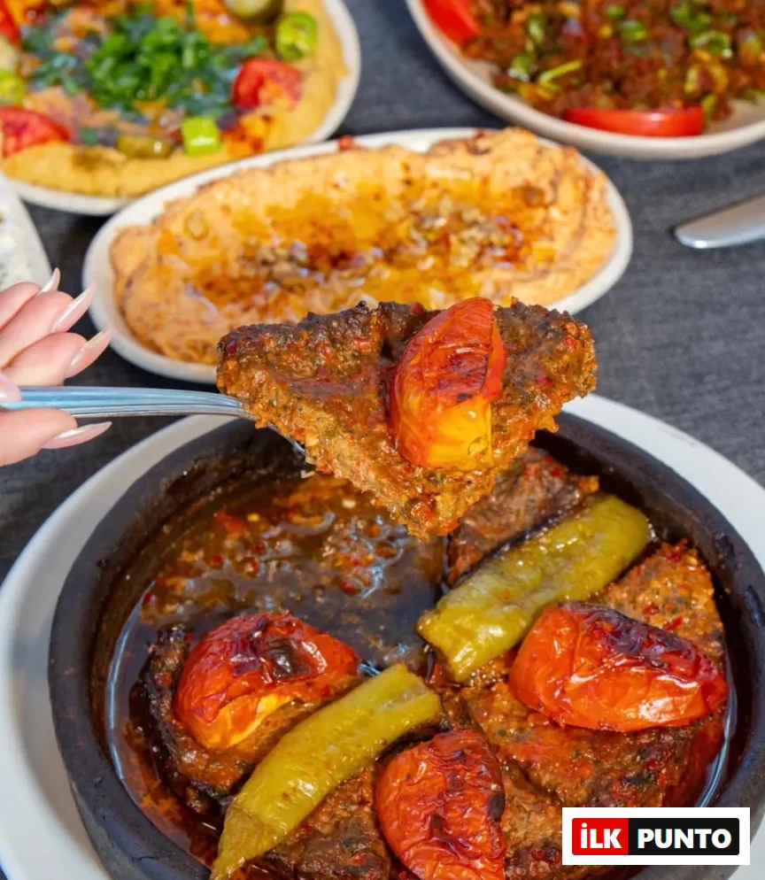 Çataldaki bir elin, kızarmış domates ve biberle zenginleştirilmiş, baharatlı Antakya tepsi kebabı benzeri et yemeğinden bir porsiyonu lezzetli sosuyla güveçten aldığı yakın çekimde, arka planda Antakya mutfağına özgü başka mezeler bulanık şekilde görülüyor.