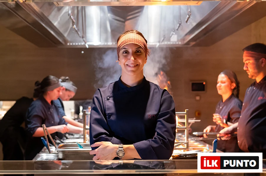 Suna Hakyemez, Roka İstanbul'un mutfağında, baş aşçı üniforması ve bandana ile kollarını kavuşturmuş, arkasında yoğun bir şekilde çalışan ekibi ve yükselen buharlar eşliğinde gülümseyerek duruyor, İstanbul’un en popüler sushi restoranları arasındaki Roka'nın deneyimli şefi olarak şehrin sushi standartlarına yön veriyor.