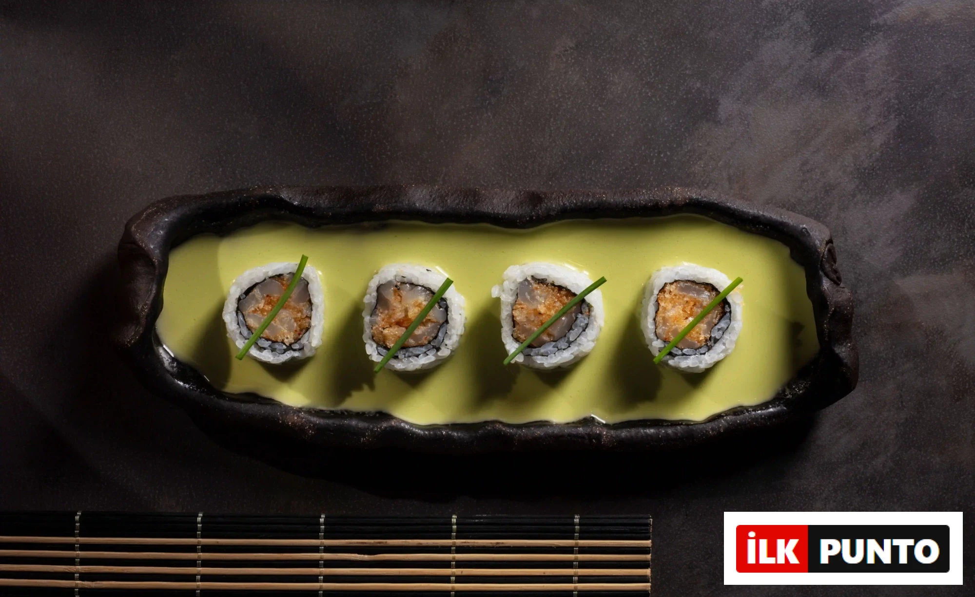 Dört adet beyaz pirinçli, ortası balık dolgulu ve taze frenk soğanı ile süslenmiş sushi rulosu, koyu renkli bir tabakta parlak sarı-yeşil bir sosun içinde özenle sunularak İstanbul’un popüler sushi restoranlarındaki şef imzalı inovatif lezzetleri yansıtıyor.
