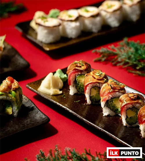 Canlı kırmızı bir zemin üzerinde, özenle hazırlanmış farklı çeşitlerdeki sushi roll'lar, yanında wasabi ve zencefil turşusu ile modern dokunuşlarla sunulmuş, İstanbul'un en popüler sushi restoranlarında tadabileceğiniz taze ve özgün lezzetleri temsil ediyor.