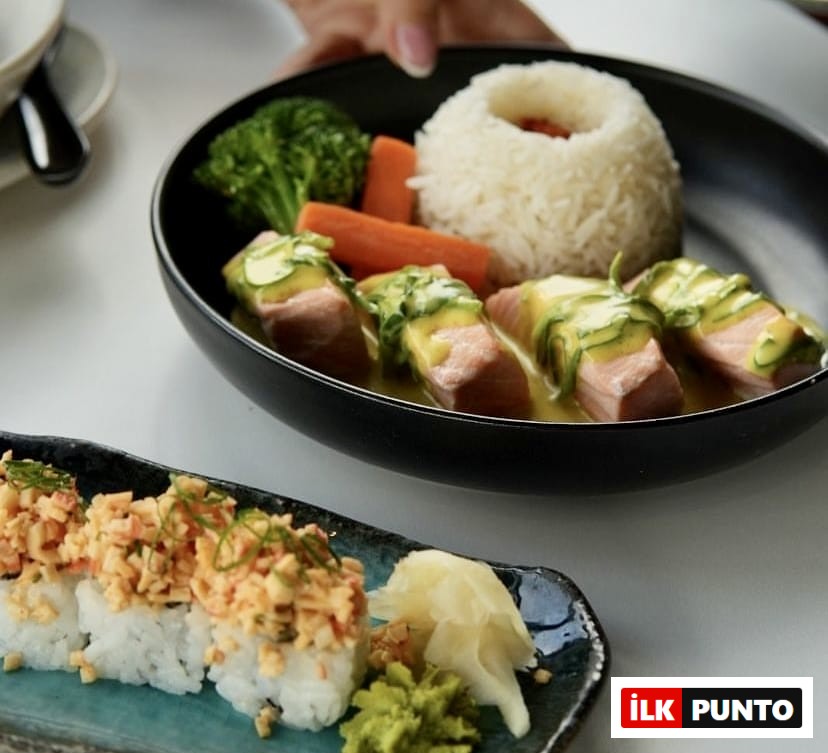 Ön planda yengeç salatası benzeri karışımla zenginleştirilmiş dört adet iştah açıcı sushi rulo, taze wasabi ve zencefil turşusu ile birlikte zarif bir tabakta sunulurken, arka planda bir elin tuttuğu siyah kasede sarı soslu ve salatalık dilimli somon filetoları, pilav, brokoli ve havuç eşliğinde, İstanbul'un en popüler sushi restoranlarının sunduğu rafine Uzak Doğu lezzetlerini ve kaliteli sunumlarını vurguluyor.