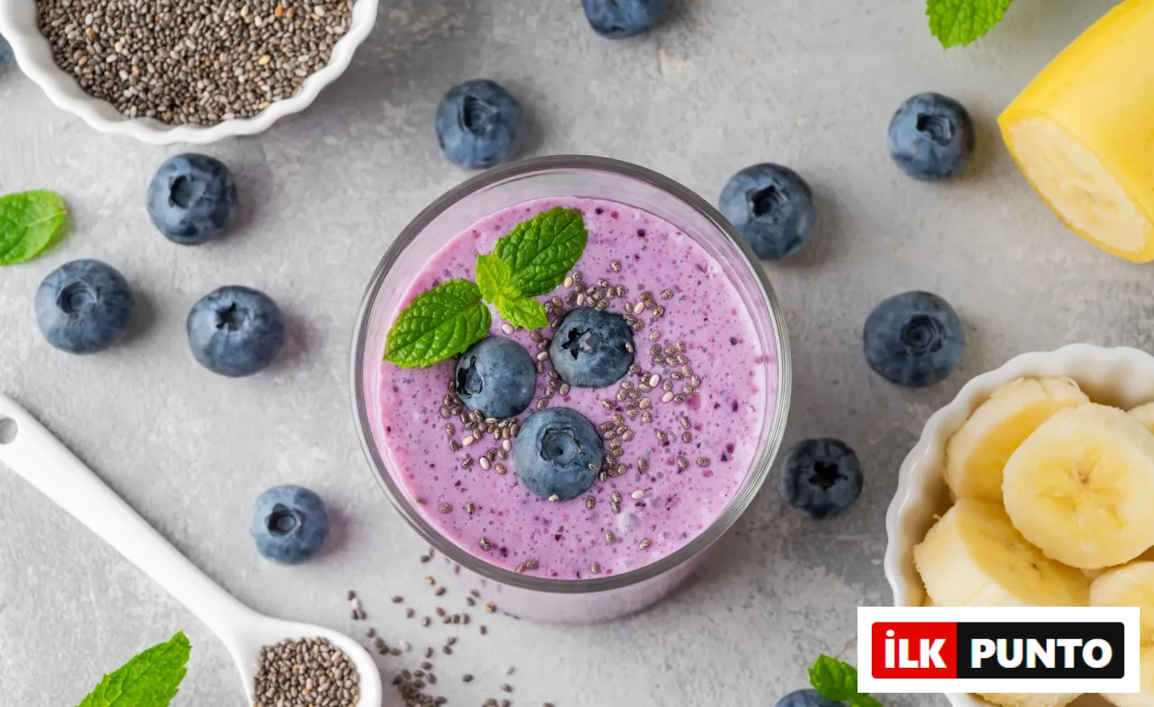 Gri bir zeminde üstten çekilmiş bu fotoğrafta, chia tohumları, yaban mersinleri ve nane yapraklarıyla süslenmiş mor renkli bir smoothie bardağı, yanında dilimlenmiş muzlar ve aronya meyvesinin cilt sağlığı için faydalarını temsil eden zengin besinleri sergiliyor.