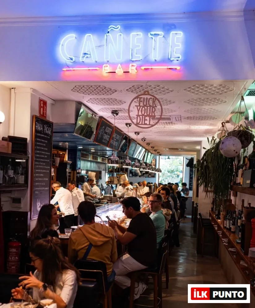 Cañete Bar'ın iç mekanı, parlak mavi neon tabelası altında, kalabalık müşterilerin bar tezgahında ve masalarda oturarak yemek yiyip sosyalleştiği hareketli bir sahneyi gösteriyor; arka plandaki açık mutfakta beyaz şef üniformalı aşçılar hazırlık yaparken, tavandaki FUCK YOUR DIET yazısı eğlenceli bir detay katıyor.