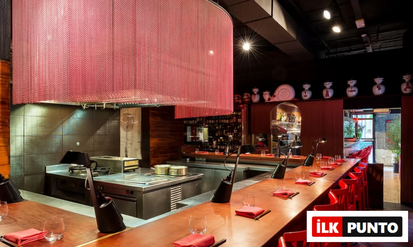 Dos Palillos restoranının modern iç mekanında, uzun ahşap tezgah boyunca sıralanmış parlak kırmızı sandalyeler, kırmızı zincirlerden oluşan dairesel bir davlumbazın altında şık bir açık mutfak alanı ve kendine özgü dekoratif objelerle bütünleşerek sıcak ve davetkar bir yemek deneyimi sunuyor.