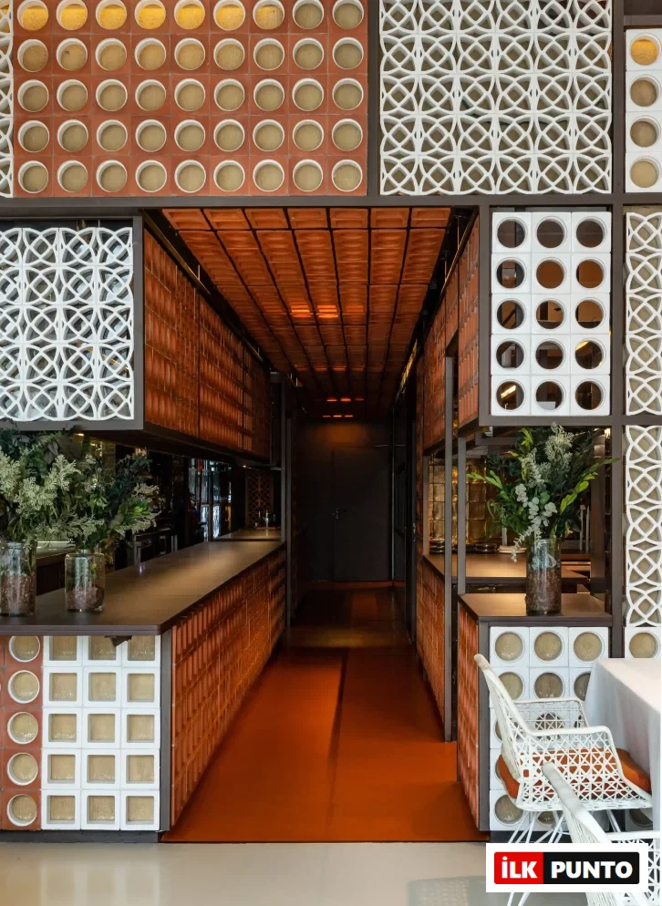 Kırmızı tuğla ve beyaz geometrik desenli panellerle süslü, sıcak turuncu zeminli uzun bir koridor ve yemek alanının görüldüğü bu modern restoran tasarımı, bitki aranjmanları ve beyaz sandalyelerle özgün bir atmosfere sahip.