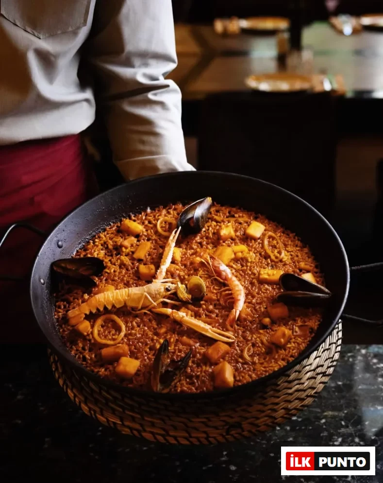 Beyaz gömlekli ve kırmızı önlüklü bir garsonun tuttuğu büyük siyah bir paella tavasında, bol miktarda karides, midye, kalamar halkaları ve belirgin bir langust ile zenginleştirilmiş kızıl-kahverengi pirinçlerden oluşan, iştah açıcı ve rengarenk deniz ürünleri paellası, hasır bir altlık üzerinde gururla sunuluyor.