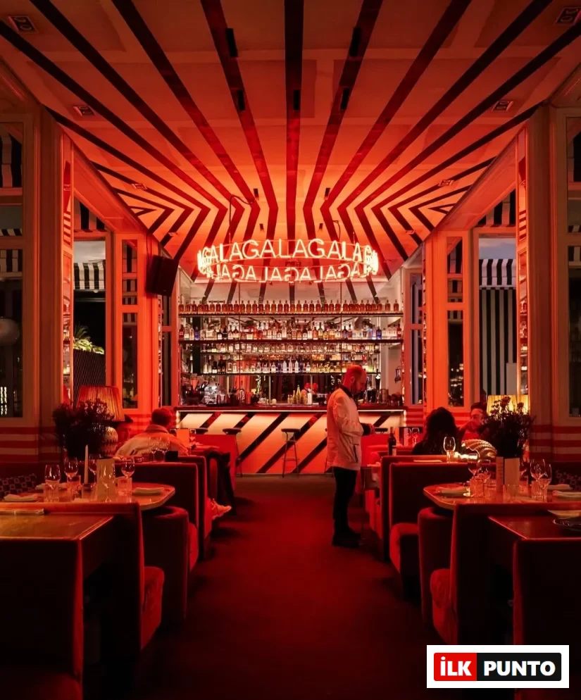 Kırmızı ışıklarla dolu, modern tasarımlı bu lüks restoranda, tavandaki çarpıcı siyah-kırmızı çizgili desenler ve barın üzerindeki parıldayan GALA neon tabelası, atmosferik bir gala gecesi veya özel etkinlik için şık bir ortam sunarken, kırmızı kadife koltuklarda oturan misafirler masalarda sosyalleşiyor.
