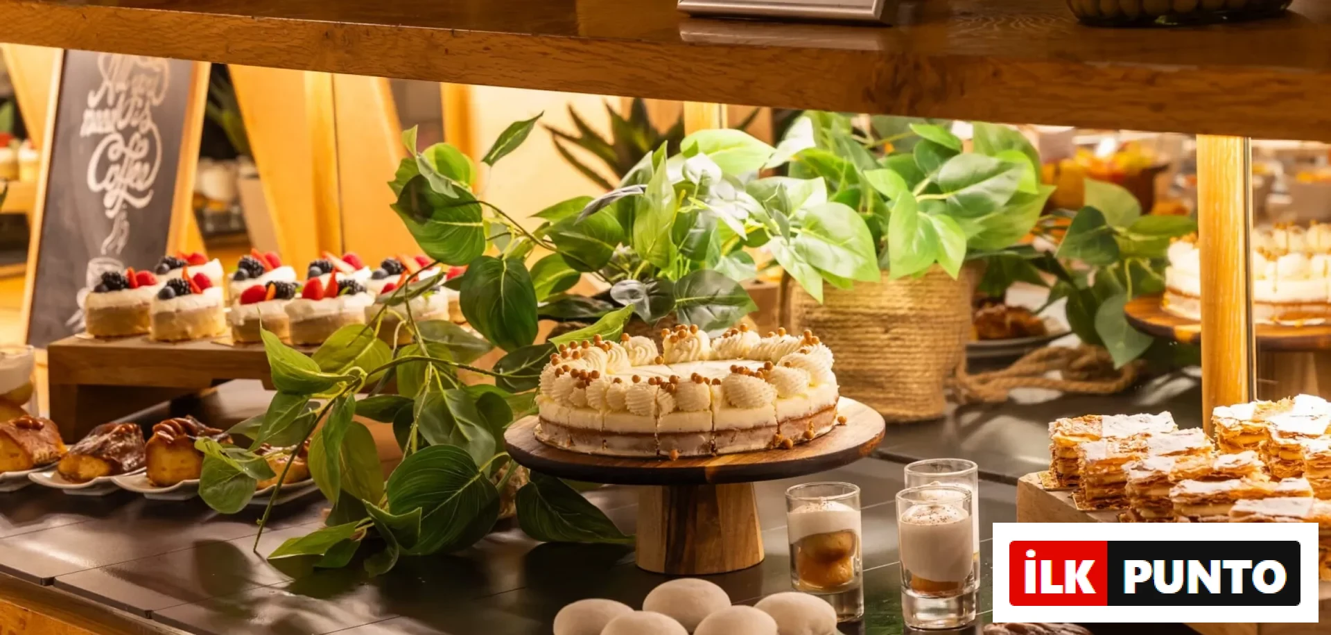 Bir otel veya restoranda özenle hazırlanmış, ortasında dilimlenmiş kremalı ve topçuk süslemeli büyük bir pastanın durduğu, yanında karışık meyveli tartletler, kat kat pudra şekerli milföy tatlıları ve yeşil bitkilerle dekore edilmiş zengin bir tatlı büfesi sunumu.