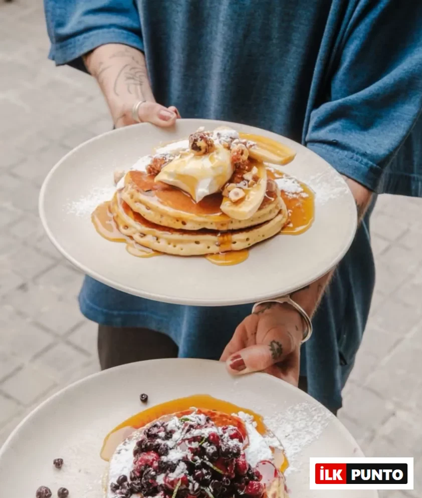 Mavi bir gömlek giyen ve dövmeleri olan bir kişinin ellerinde, üstte muz dilimleri, çırpılmış krema ve fındıklarla süslenmiş karamel soslu üç kat pancake, altta ise böğürtlen ve frambuaz gibi çeşitli meyvelerle zenginleştirilmiş, pudra şekeri serpilmiş bir diğer lezzetli pancake tabağı, brunch veya kahvaltı keyfi için sunuluyor.