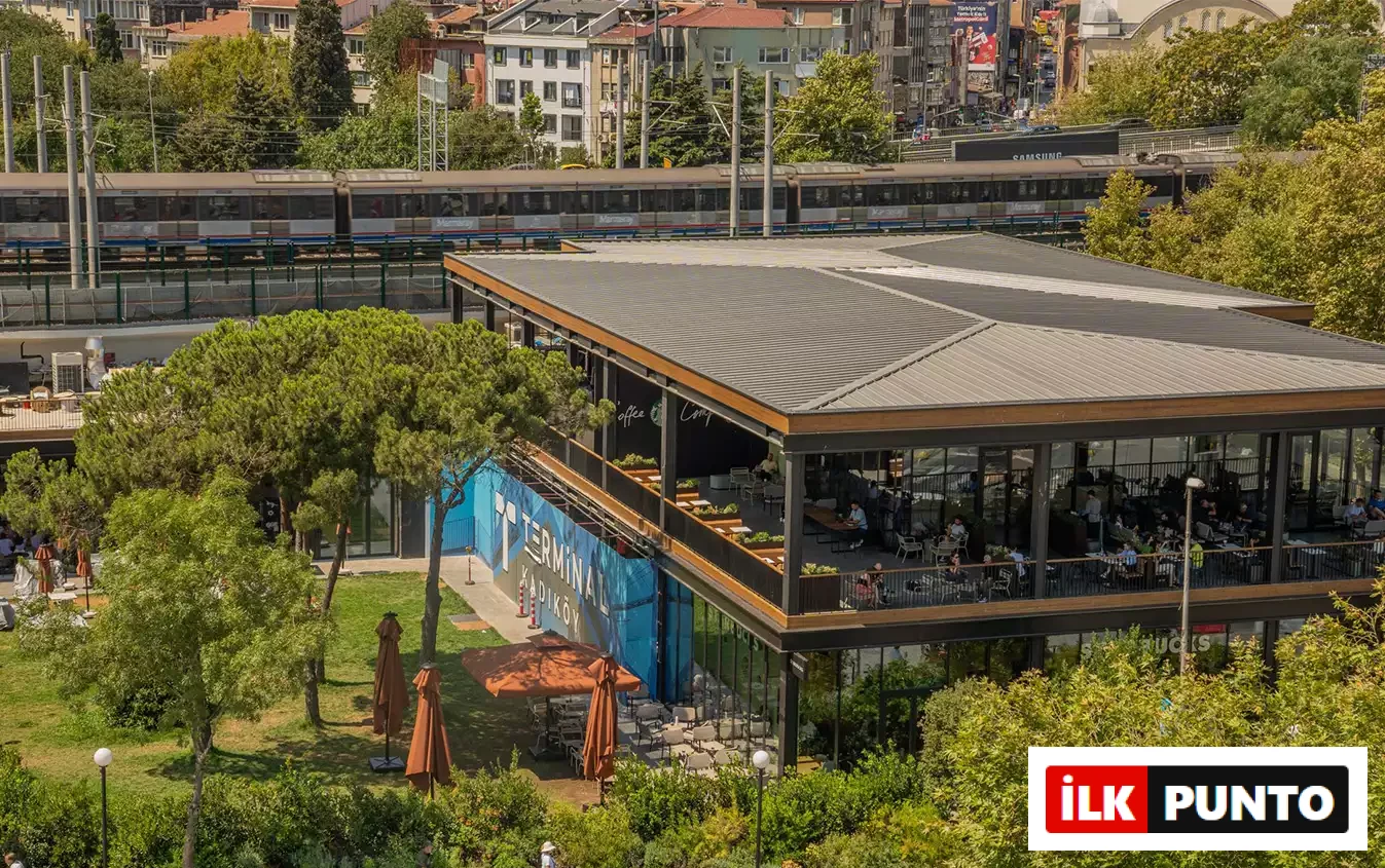 İlk Punto - Terminal Kadıköy Rehberi: Lezzet, Alışveriş ve Eğlence Durakları. Arka planda geçen Marmaray treninin hemen yanında, yeşil alanlarla çevrili iki katlı cam mimarili Terminal Kadıköy binası. Ziyaretçiler yeni yaşam alanının açık hava teraslarında sosyalleşiyor.