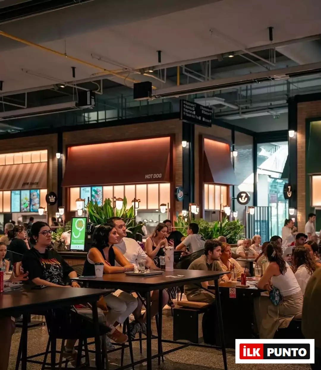 İlk Punto Terminal Kadıköy Rehberi: Lezzet, Alışveriş ve Eğlence Durakları. Terminal Kadıköy'ün geniş 7DE7 yeme-içme alanında yüksek masalarda oturup sohbet eden ve yemek yiyen ziyaretçiler. Arka planda Hot Dog standı, bitkiler ve sıcak aydınlatmalarla modern endüstriyel gastronomi atmosferi.