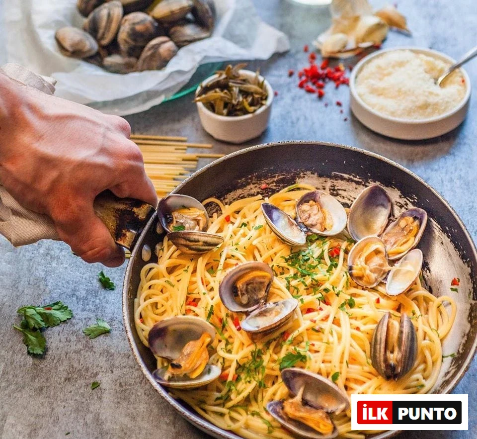 Bir elin tavayı tuttuğu karede, üzerinde taze istiridyeler, doğranmış yeşillikler ve kırmızı biber parçacıklarıyla süslenmiş lezzetli spaghetti vongole tabağı Selanik'in en iyi restoranlarında bulunabilecek zengin deniz ürünleri seçeneklerini temsil ediyor, arka planda çiğ istiridyeler, turşular ve rendelenmiş peynir görülüyor.