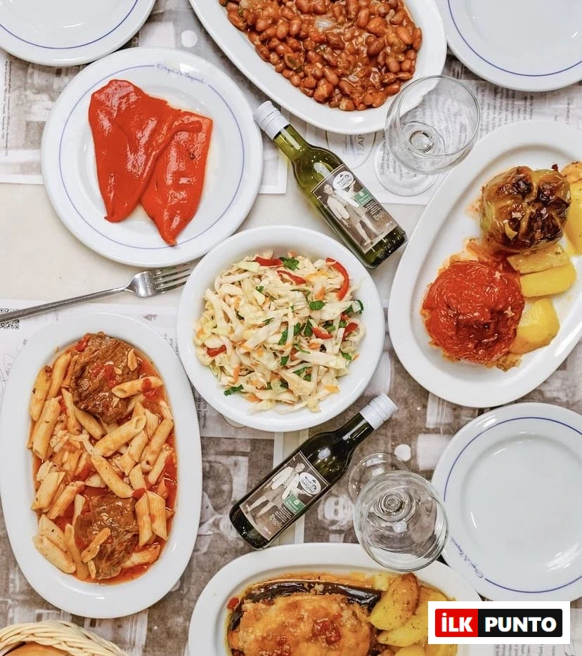 Selanik'teki geleneksel bir tavernada sunulan; kavrulmuş kırmızı biberler, kuru fasulye pilaki, lahana salatası, etli penne makarna, domates ve biber dolmaları ile patatesli patlıcan yemeği gibi çeşitli otantik Yunan lezzetleri ve iki şişe beyaz şarapla dolu, tepeden çekilmiş zengin bir masa görünümü.