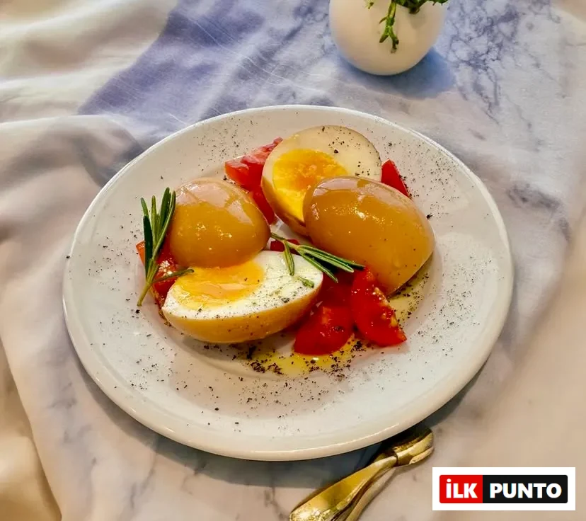 Beyaz bir tabakta, geleneksel Selanik yumurtası veya zeme yumurtası olarak bilinen, dışı kararmış ve içi kremsi sarı renkte pişmiş haşlanmış yumurtalar özenle yerleştirilmiş. Yumurtaların bazıları ikiye bölünmüş olup, hafif akışkan parlak sarı yumurta sarısı belirgin bir şekilde görülüyor. Tabağın etrafında taze doğranmış kırmızı domatesler, aromatik taze biberiye dalları, zeytinyağı ve karabiber serpilmiş; bu iştah açıcı sunum kahvaltı sofraları için ideal.