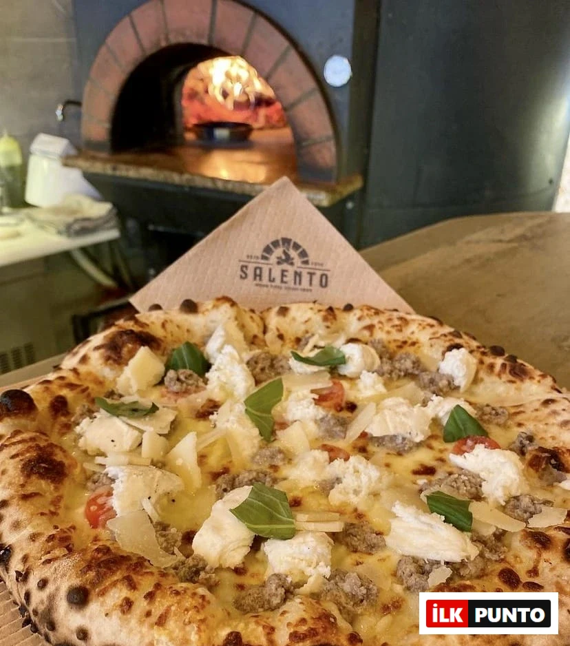 Selanik'teki Salento'dan servis edilmiş, kenarları hafifçe kömürleşmiş, üzerinde bol sosis, kremsi ricotta, rendelenmiş parmesan peyniri, domates parçacıkları ve taze fesleğen yaprakları bulunan, arka planda alevli taş fırının görüldüğü ağız sulandırıcı bir pizza.