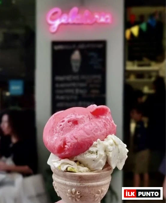 Elinde, üstünde pembe bir top ve altında vanilya veya fıstık dondurması olan, desenli bir külahı tutan kişinin yakın çekim fotoğrafında, arka planda pembe neon Gelateria tabelası ve bulanık bir dondurmacı dükkanı görülüyor.